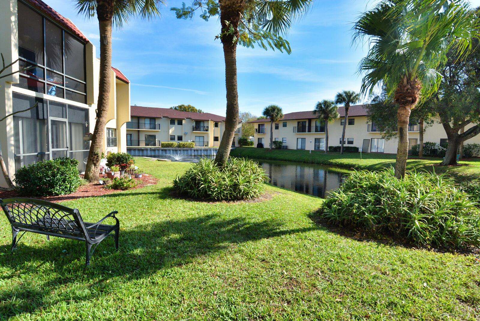 A black metal bench sits on a grassy lawn beside a pond and apartment buildings — 2050 Oleander Boulevard # 306, Fort Pierce FL 34950, FORT PIERCE, FL