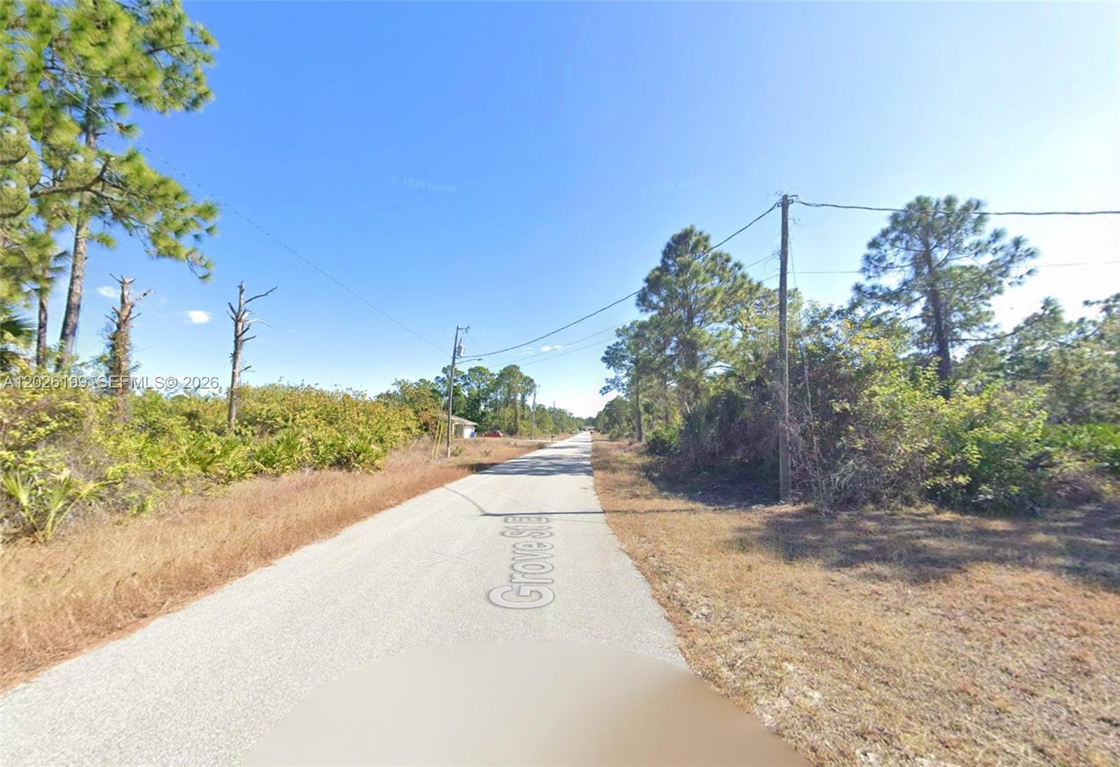 A paved road stretches into the distance, flanked by trees and brush under a clear sky — 1047 Grove St E, Lehigh Acres FL 33974, LEHIGH ACRES, FL