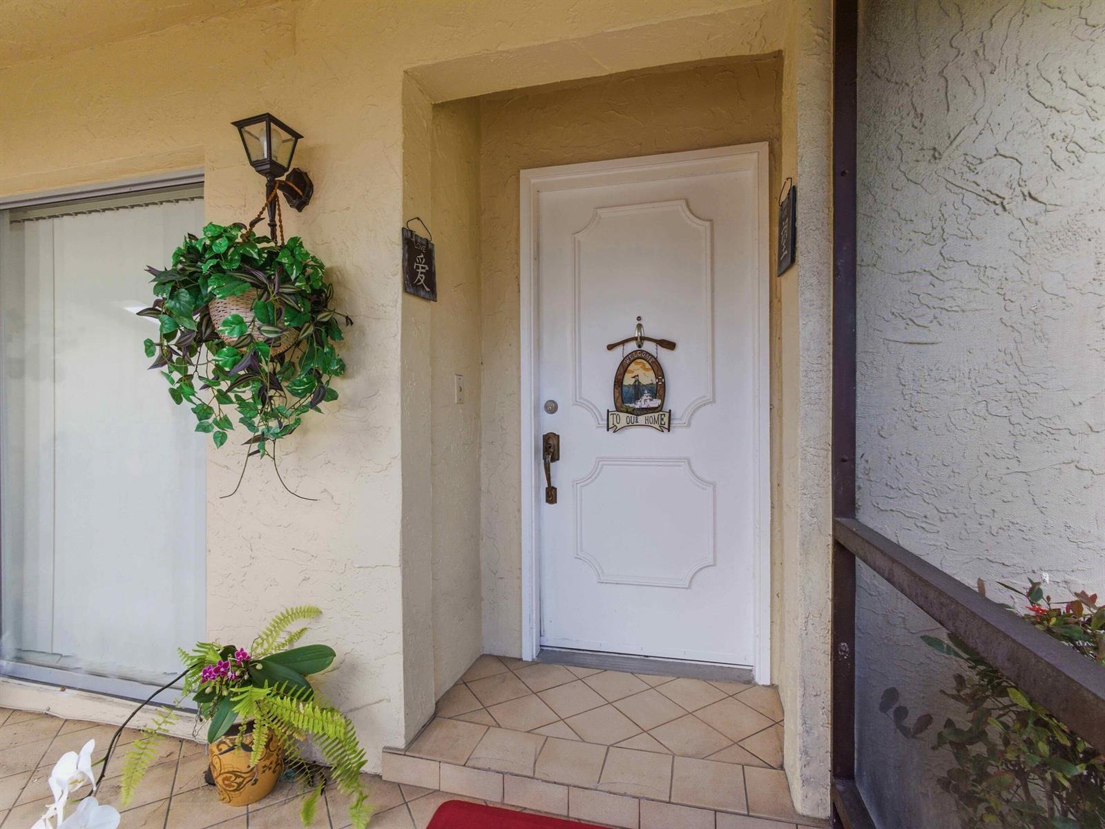 A white front door with a welcome sign sits on a tiled porch with plants — 13773 Via Aurora # C, Delray Beach FL 33484, DELRAY BEACH, FL