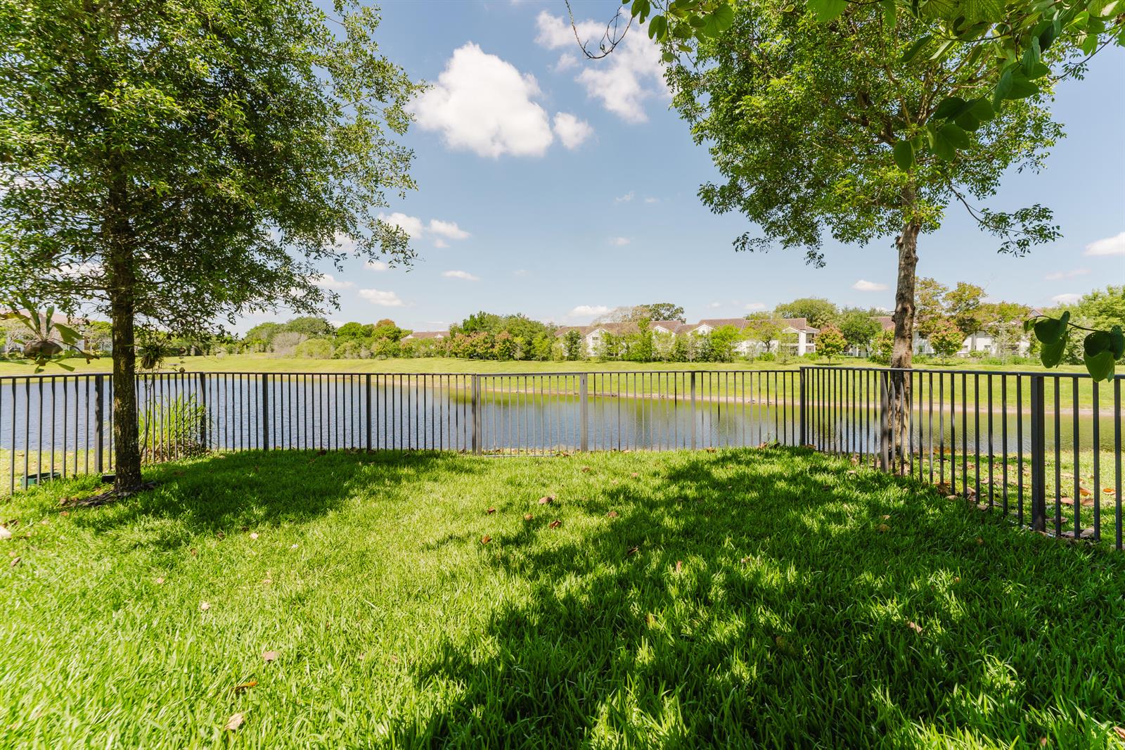 A grassy backyard features a metal fence overlooking a pond with trees and distant buildings — 295 NW 33rd Lane, Pompano Beach FL 33069, POMPANO BEACH, FL