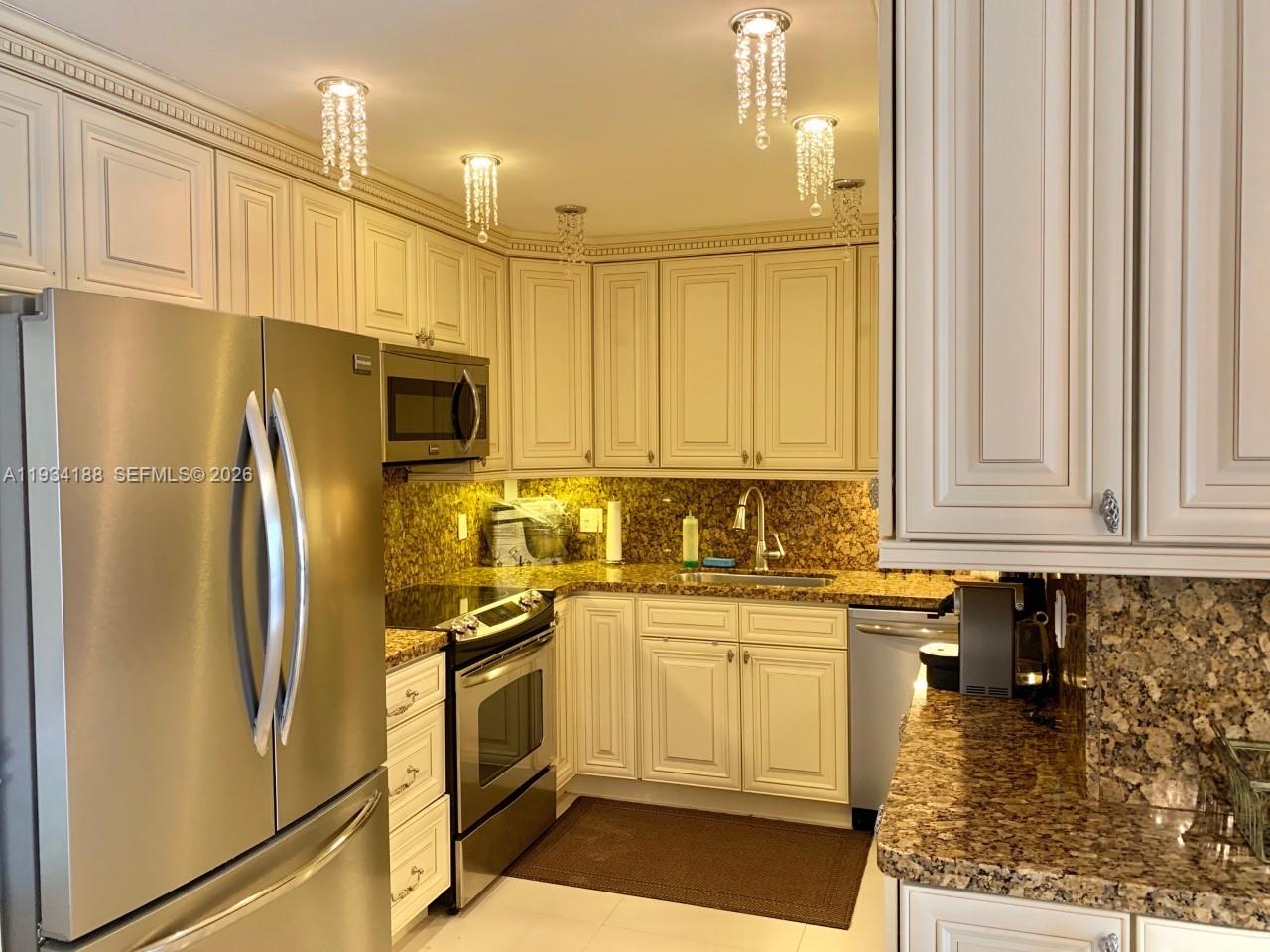 A kitchen features off-white cabinets, granite countertops, stainless steel appliances, and crystal pendant lighting — 1985 S Ocean Dr # 3C, Hallandale Beach FL 33009, HALLANDALE BEACH, FL