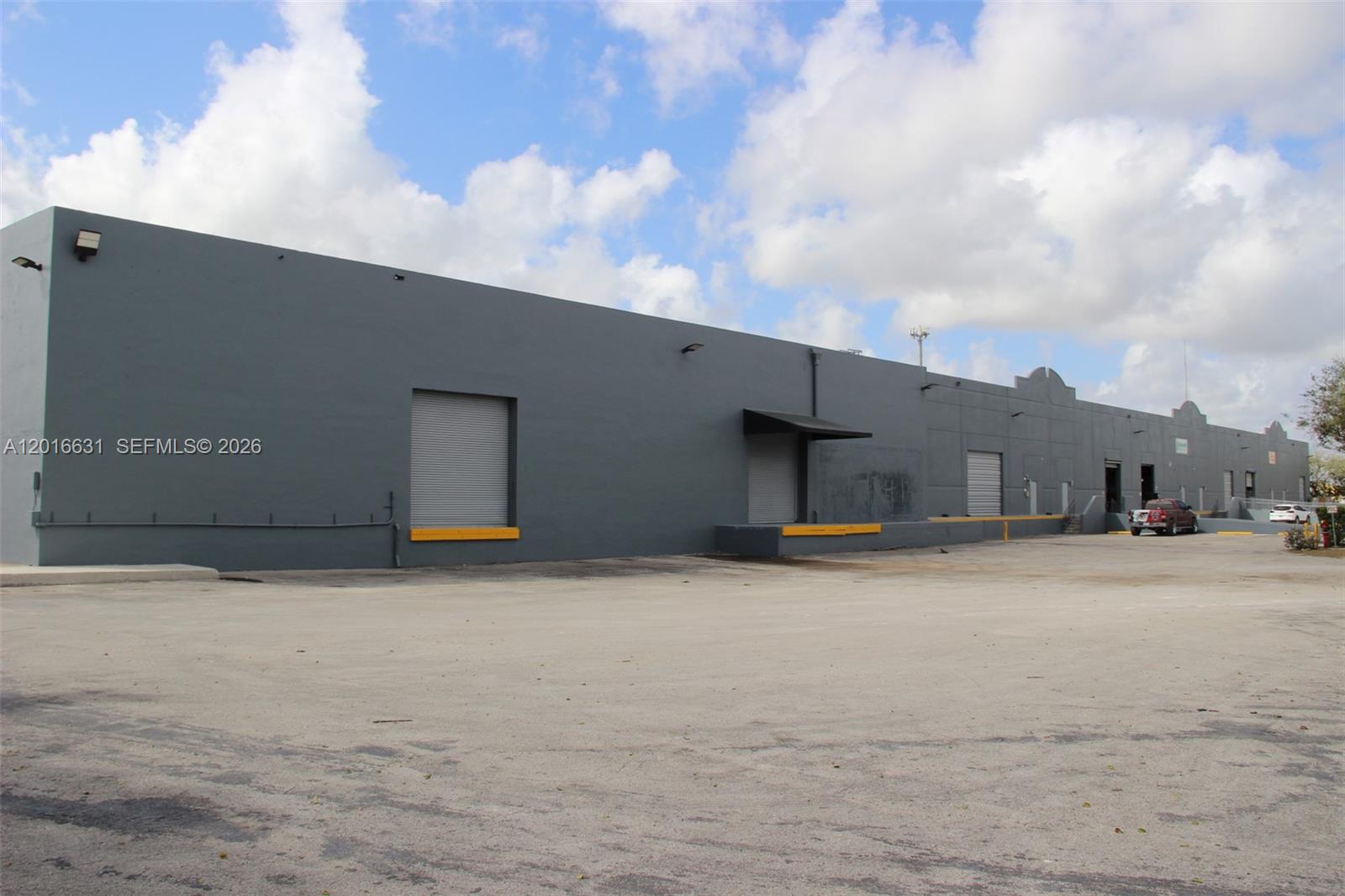 A long, gray industrial warehouse building features multiple loading dock doors and a paved lot — HOMESTEAD, FL
