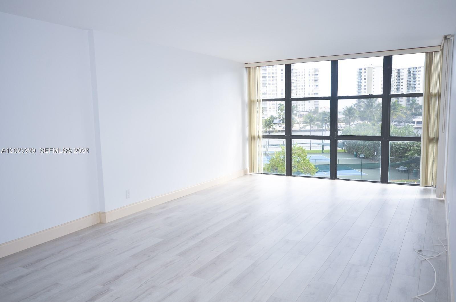An empty room features light wood-look flooring, white walls, and floor-to-ceiling windows overlooking tennis courts — 1000 Parkview Dr # 426, Hallandale Beach FL 33009, HALLANDALE BEACH, FL