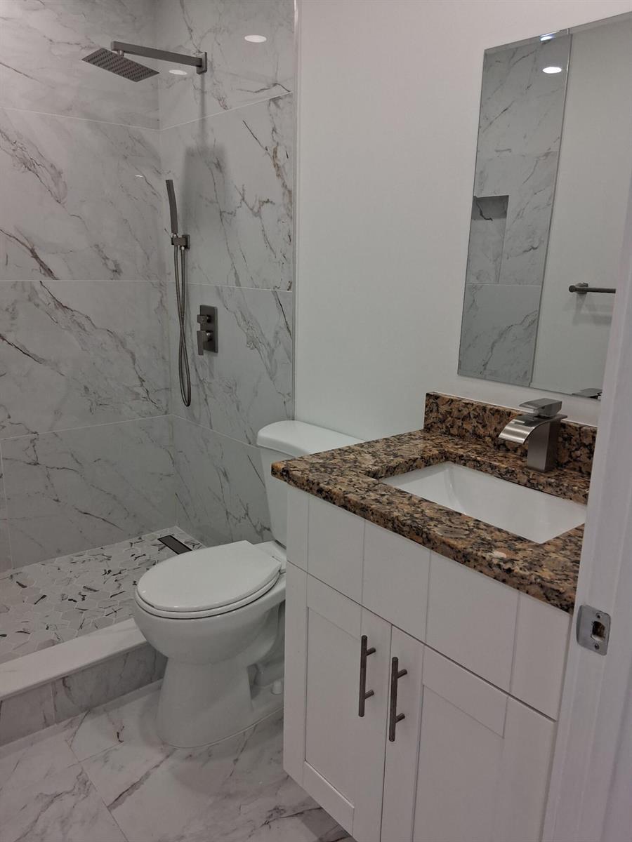 A small bathroom features a marble-tiled walk-in shower, a white vanity with granite countertop, and toilet — 2100 Springdale Boulevard # 303, Palm Springs FL 33461, PALM SPRINGS, FL