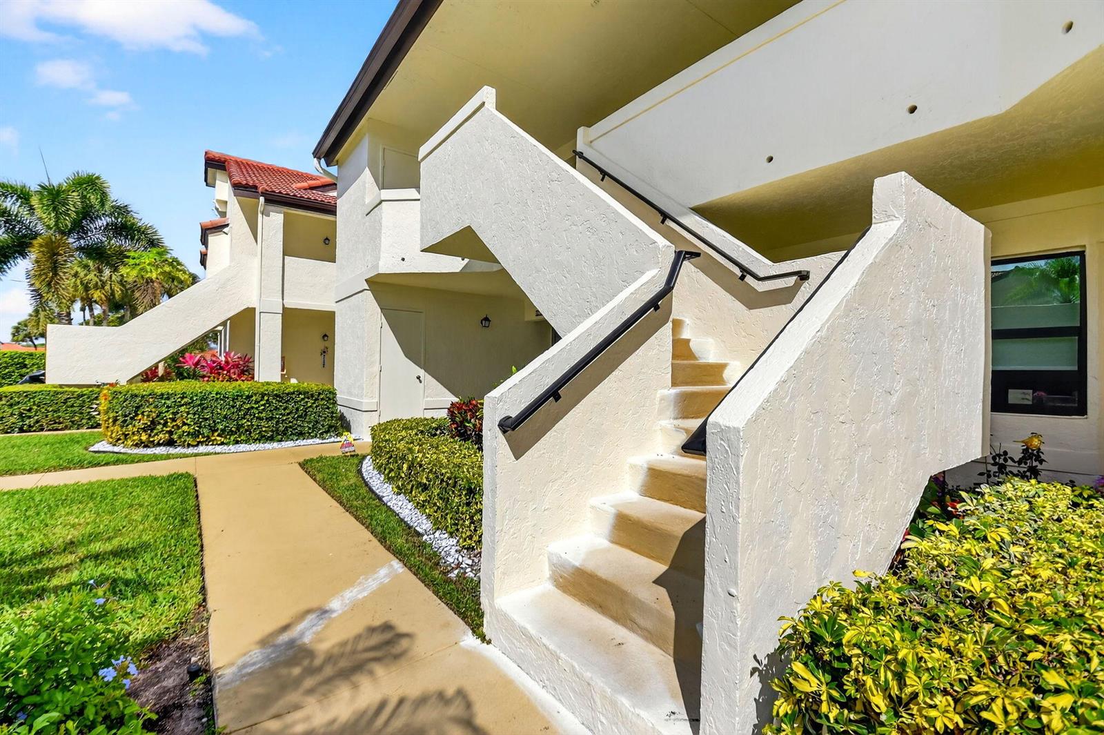 An exterior view of a multi-story apartment building featuring concrete stairs and landscaped gardens — 5903 Parkwalk Drive # 623, Boynton Beach FL 33472, BOYNTON BEACH, FL