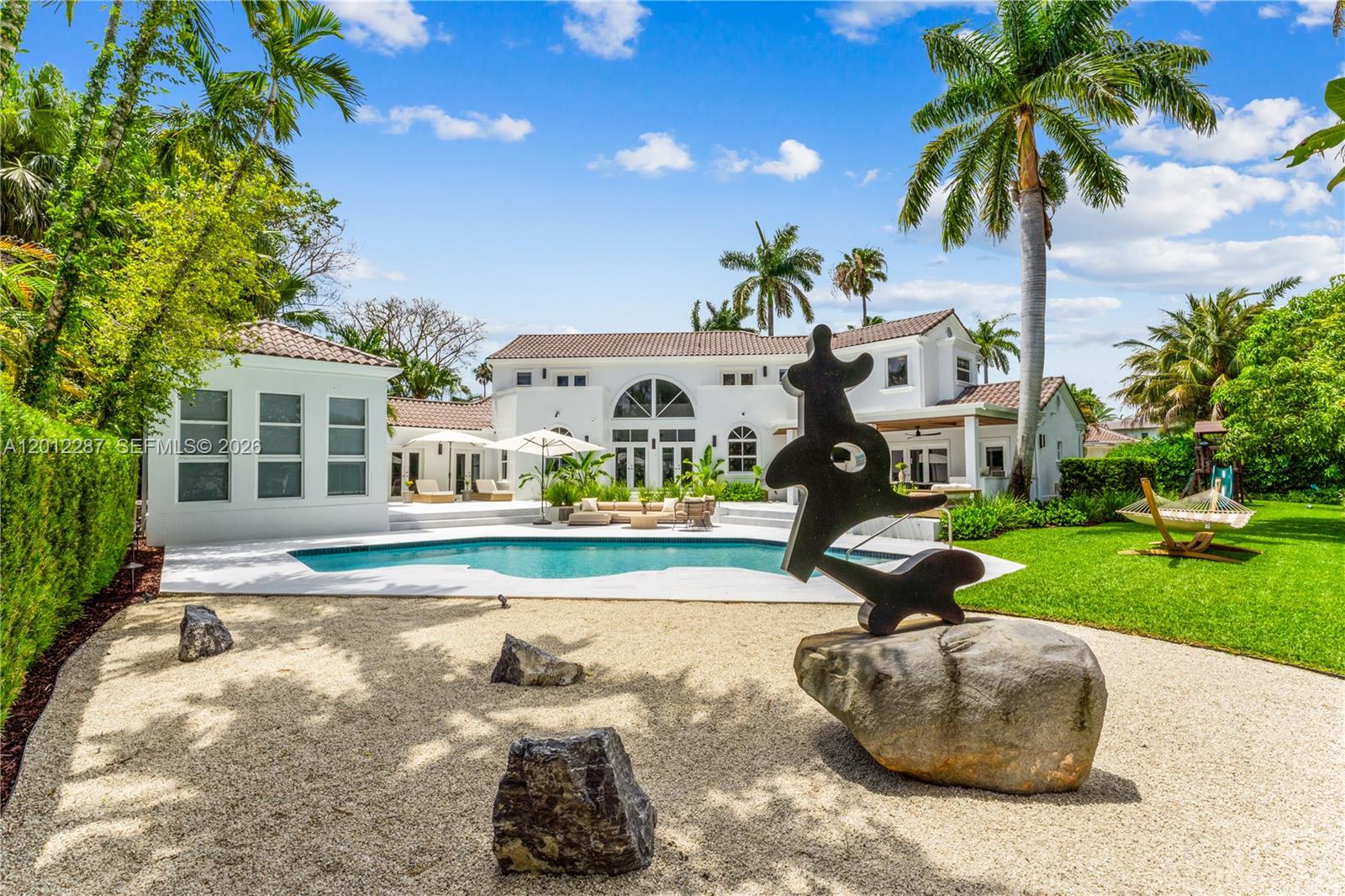 A backyard features a swimming pool, a modern sculpture on a rock, and a white house — 8763 SW 61st Pl, Pinecrest FL 33156, PINECREST, FL