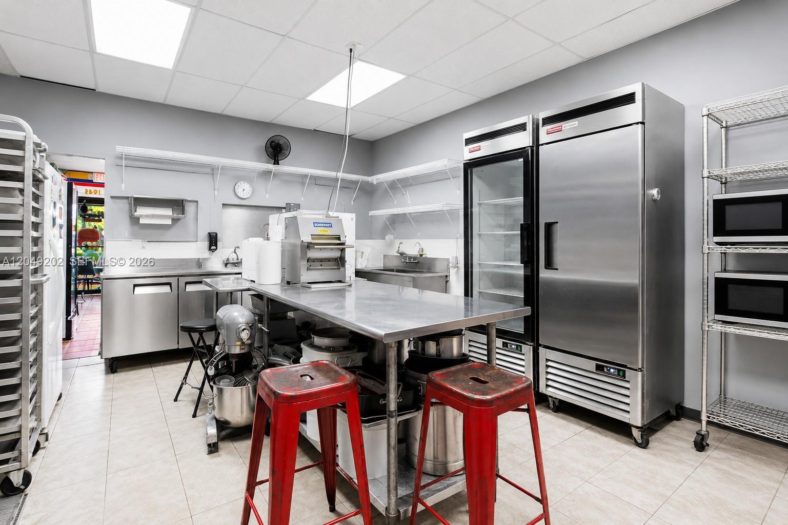 A commercial kitchen features stainless steel prep tables, appliances, and two red metal stools — HIALEAH, FL