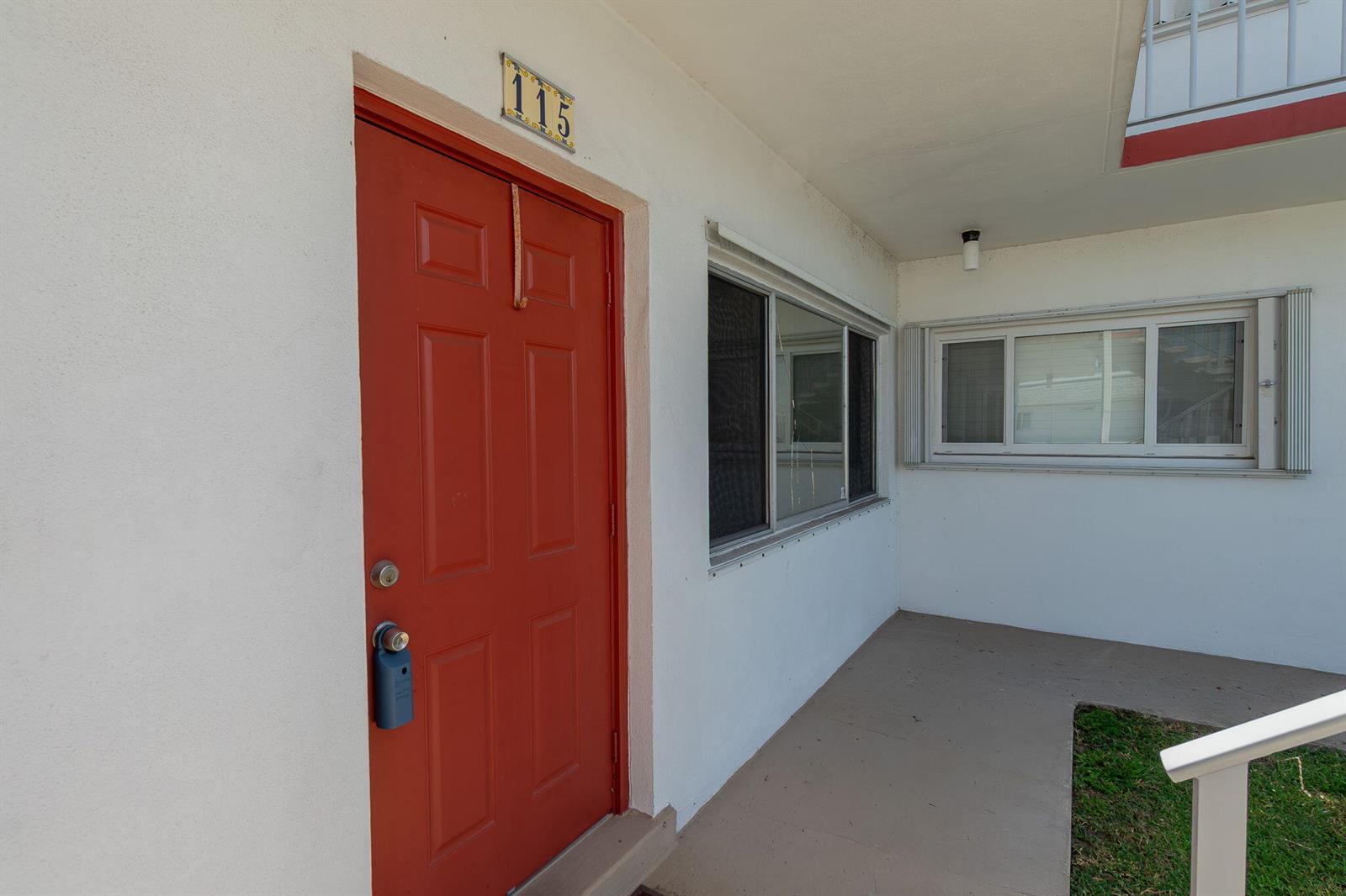 A red door with a lockbox and the number 115 sits on a white exterior wall — 3120 Cynthia Lane # 115, FL 33461, FL
