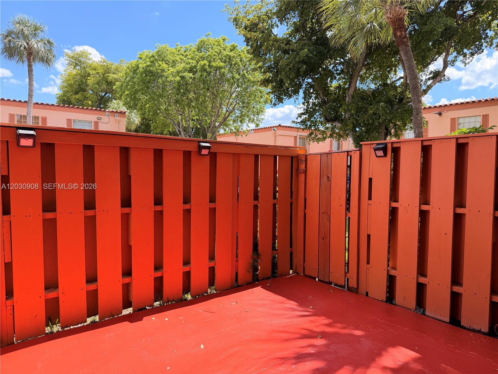 A red-painted patio is enclosed by a tall, orange wooden privacy fence with outdoor lights — MIAMI LAKES, FL