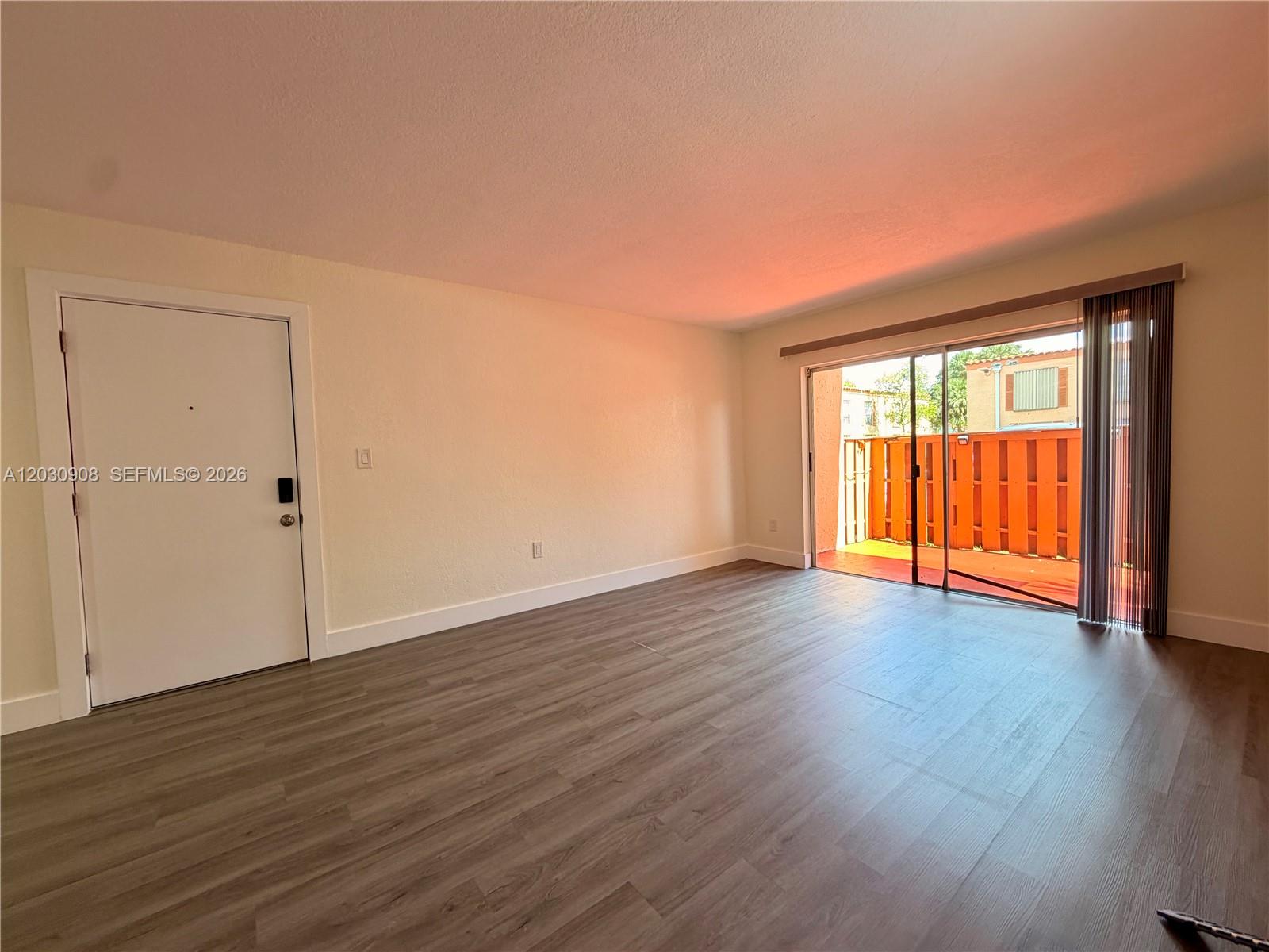 An empty room features wood-look flooring, a white entry door, and sliding glass doors to a balcony — MIAMI LAKES, FL