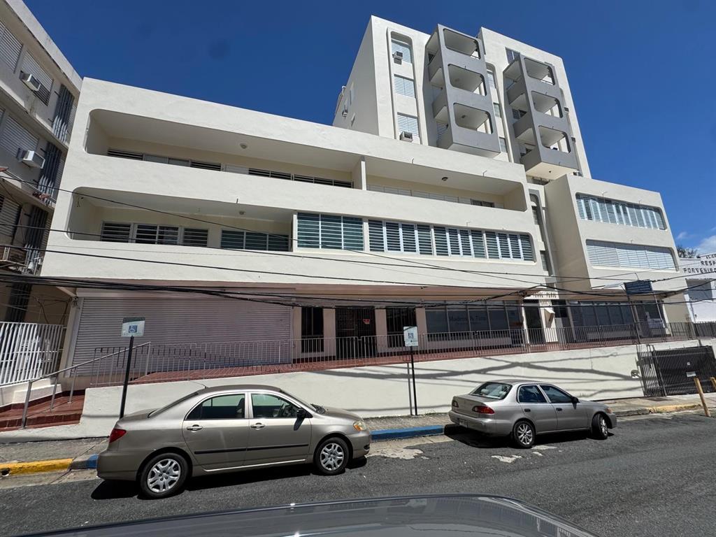 A multi-story white apartment building with balconies and a ground-level commercial space with parked cars — 17 NC PERAL UNIT 1B, MAYAGUEZ, PR
