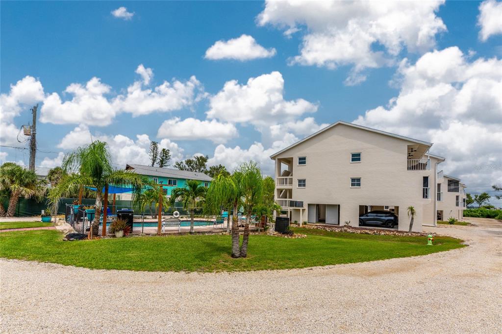 A multi-story beige building sits next to a fenced swimming pool and a gravel driveway — 5031 N BEACH ROAD UNIT 222, ENGLEWOOD, FL