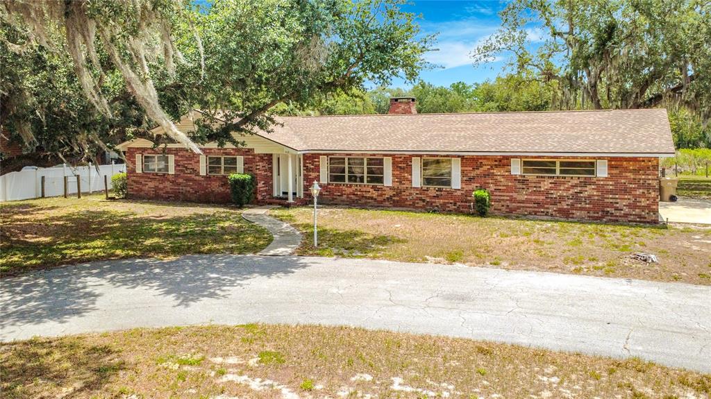 A single-story brick house with a shingled roof sits behind a curved paved driveway — 1112 CABALLO ROAD, LEESBURG, FL
