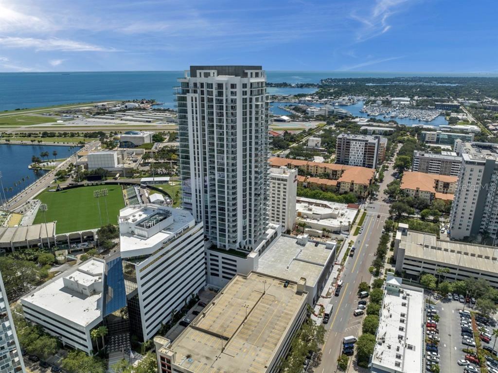 An aerial view shows a tall residential skyscraper surrounded by city buildings, parking, and water — 301 1ST STREET S UNIT 1704, ST PETERSBURG, FL
