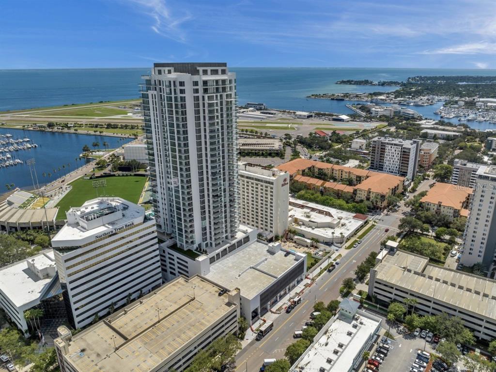 An aerial view of a tall residential tower surrounded by city buildings, parking, and water — 301 1ST STREET S UNIT 1704, ST PETERSBURG, FL