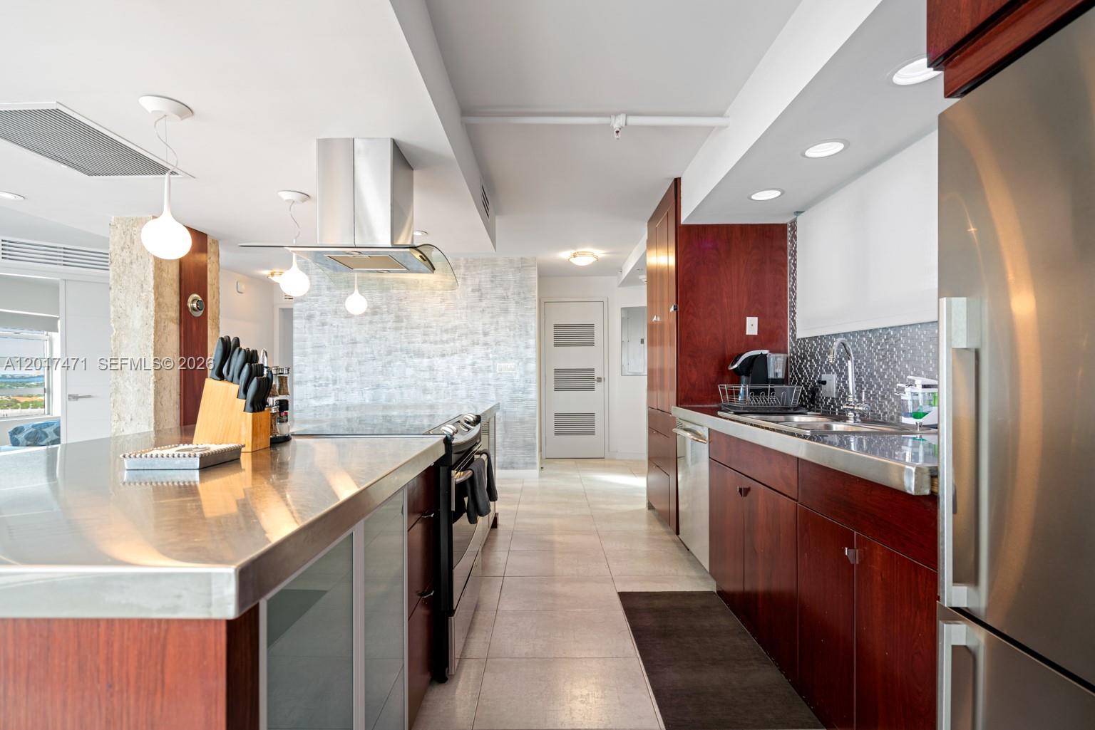 A modern kitchen features stainless steel countertops, dark wood cabinetry, and a stainless steel range hood — MIAMI BEACH, FL