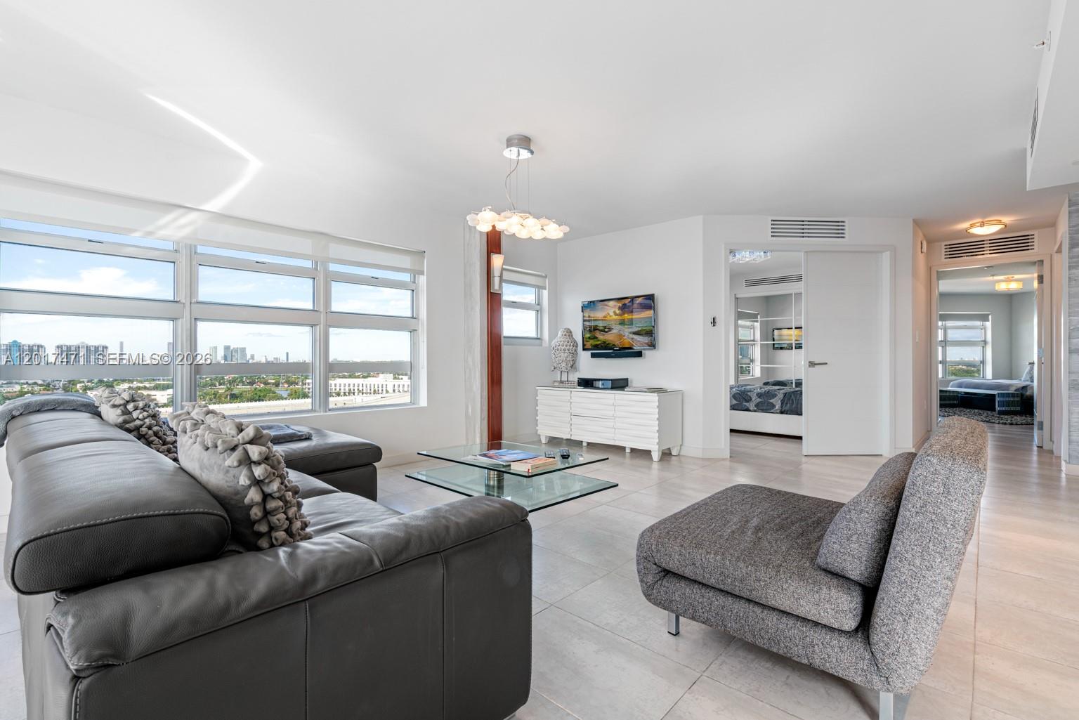 A living room features a grey leather sectional, a glass coffee table, and large windows — MIAMI BEACH, FL