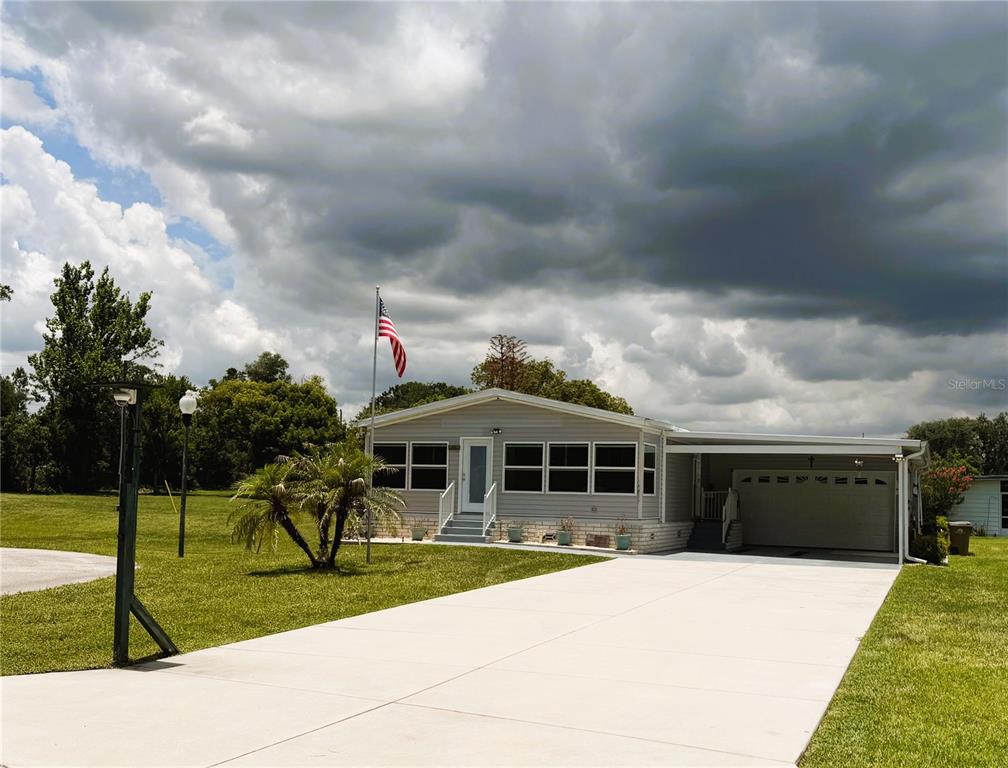 A single-story manufactured home with a carport, concrete driveway, and American flag in the yard — 165 SAGO PALM DRIVE, LEESBURG, FL