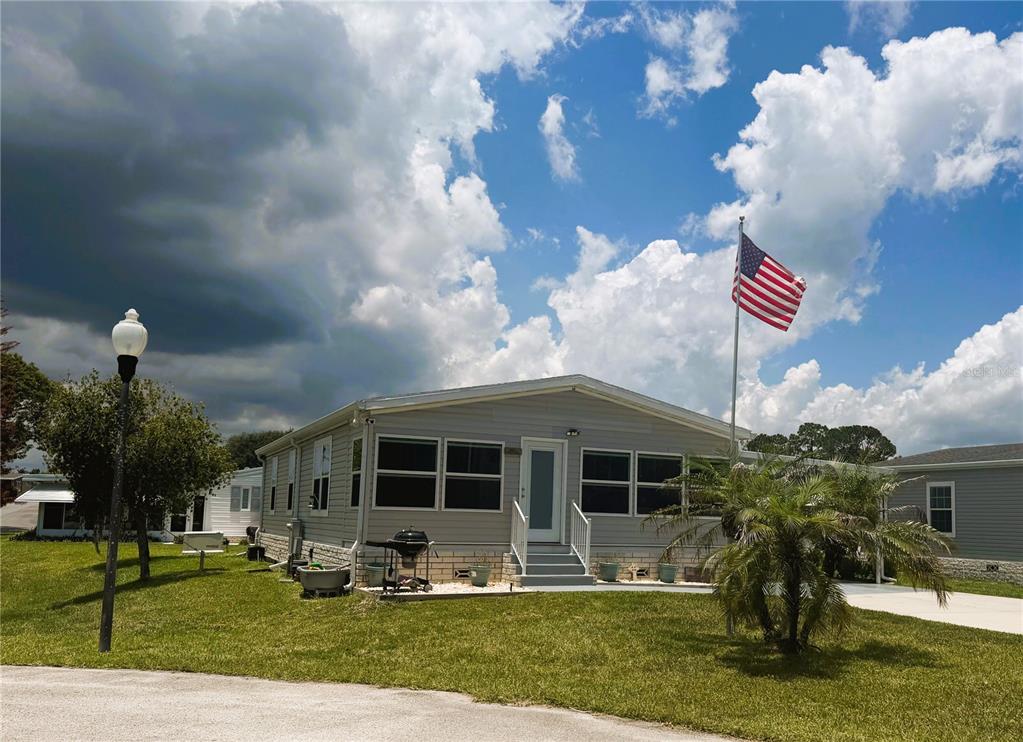 A single-story manufactured home with light gray siding and an American flag in the yard — 165 SAGO PALM DRIVE, LEESBURG, FL