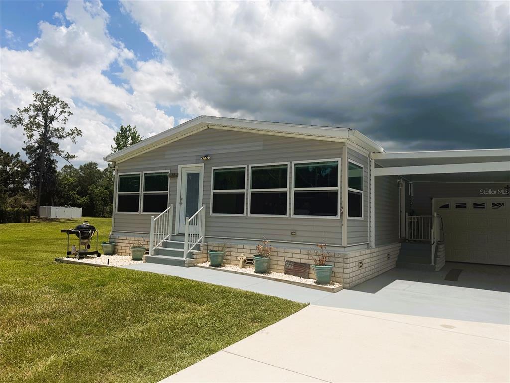A light gray manufactured home features a front porch, brick skirting, and an attached carport — 165 SAGO PALM DRIVE, LEESBURG, FL