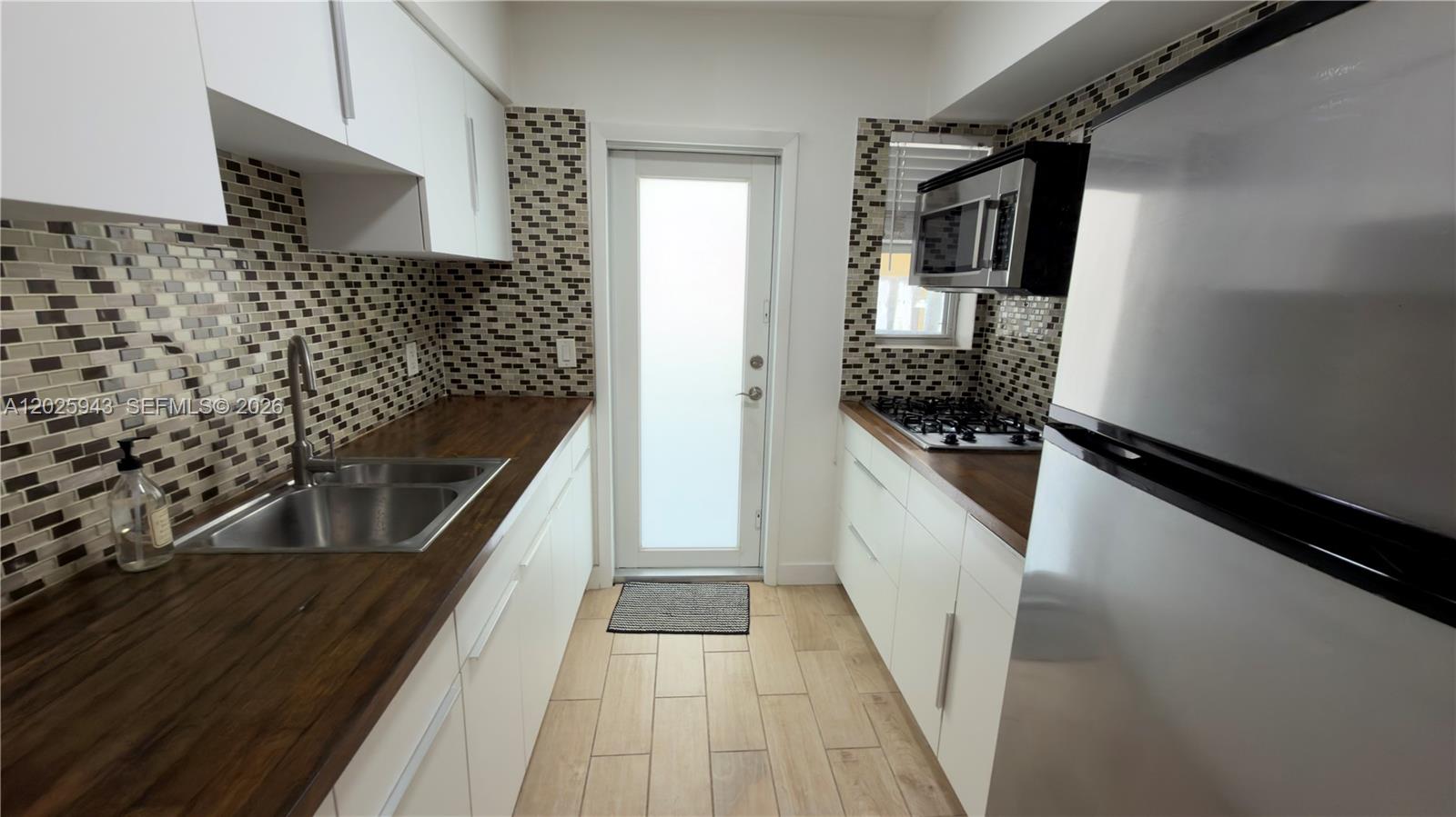 A galley kitchen features white cabinets, wood countertops, mosaic tile backsplashes, and stainless steel appliances — MIAMI BEACH, FL