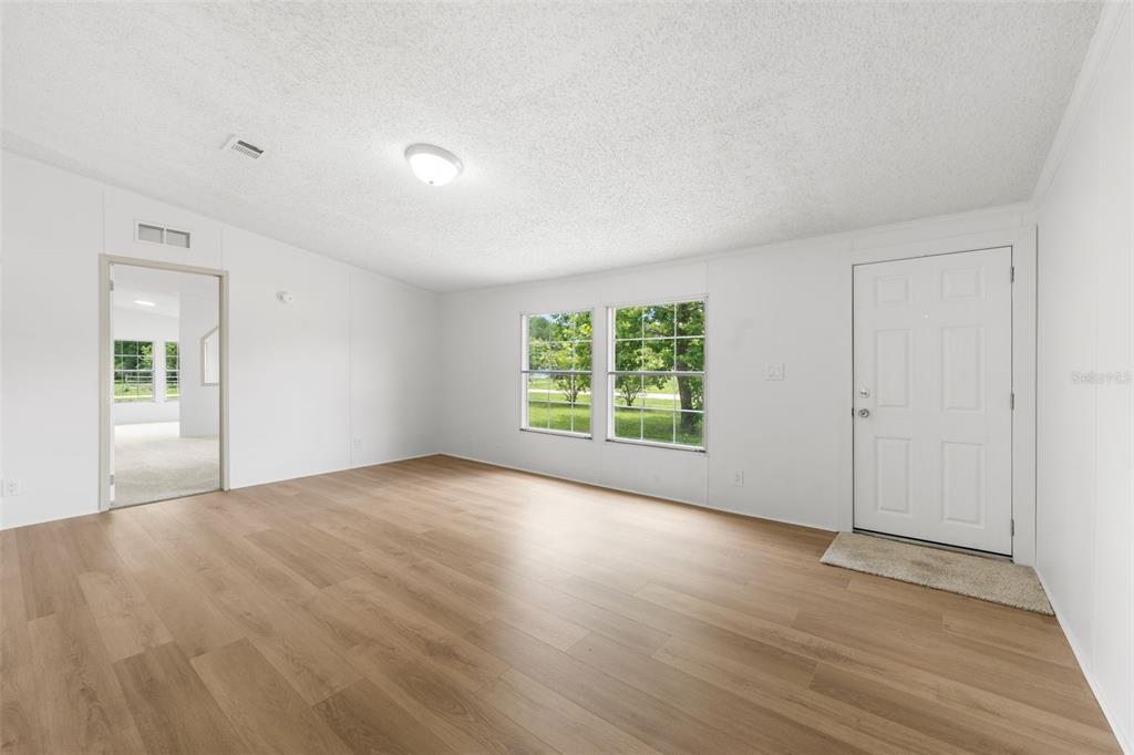 A bright, empty living room features white walls, wood-look flooring, and a white entry door — 15591 NE 46TH STREET, WILLISTON, FL
