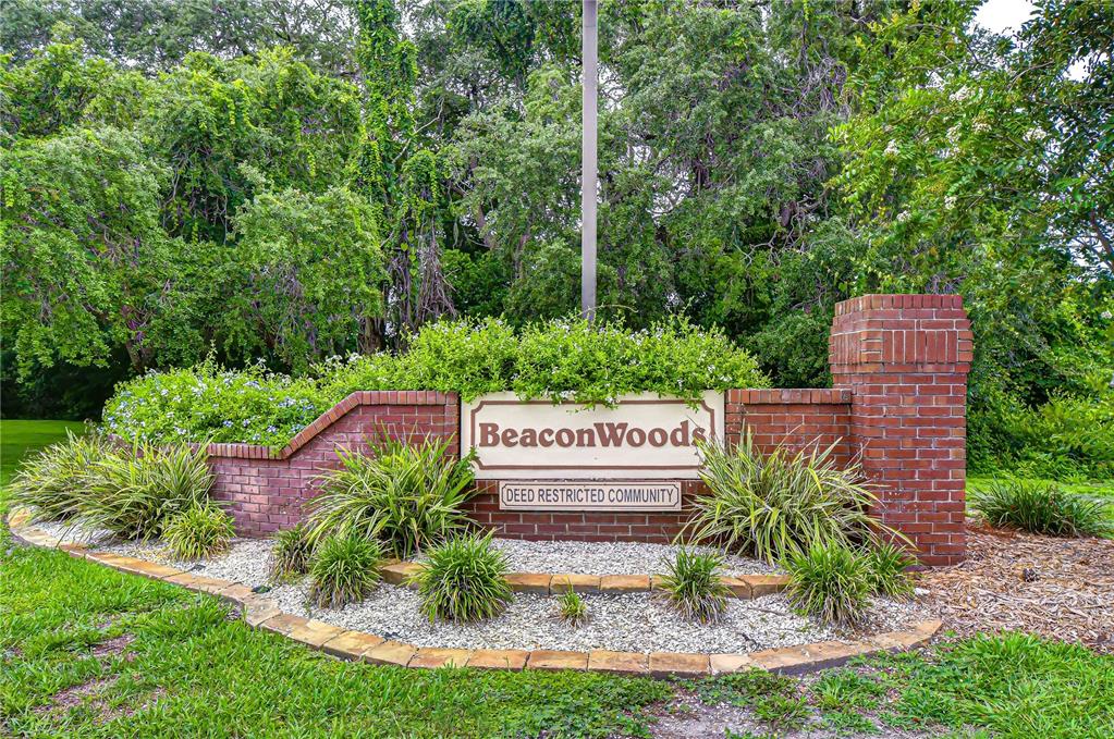 A brick monument sign for Beacon Woods stands in a landscaped garden with lush trees — 8804 VILLAGE MILL ROW, HUDSON, FL