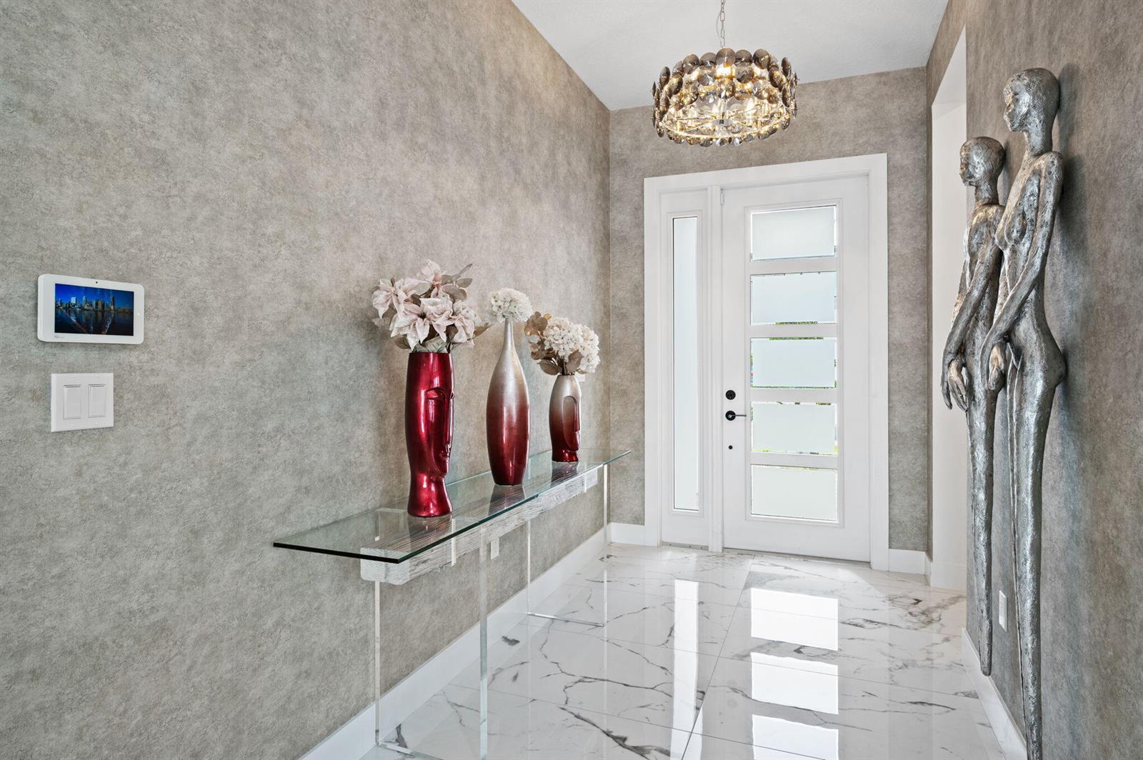 A modern entryway features marble floors, textured wallpaper, a glass console table, and metallic sculptures — 8599 Tower Bridge Court, Boca Raton FL 33496, BOCA RATON, FL