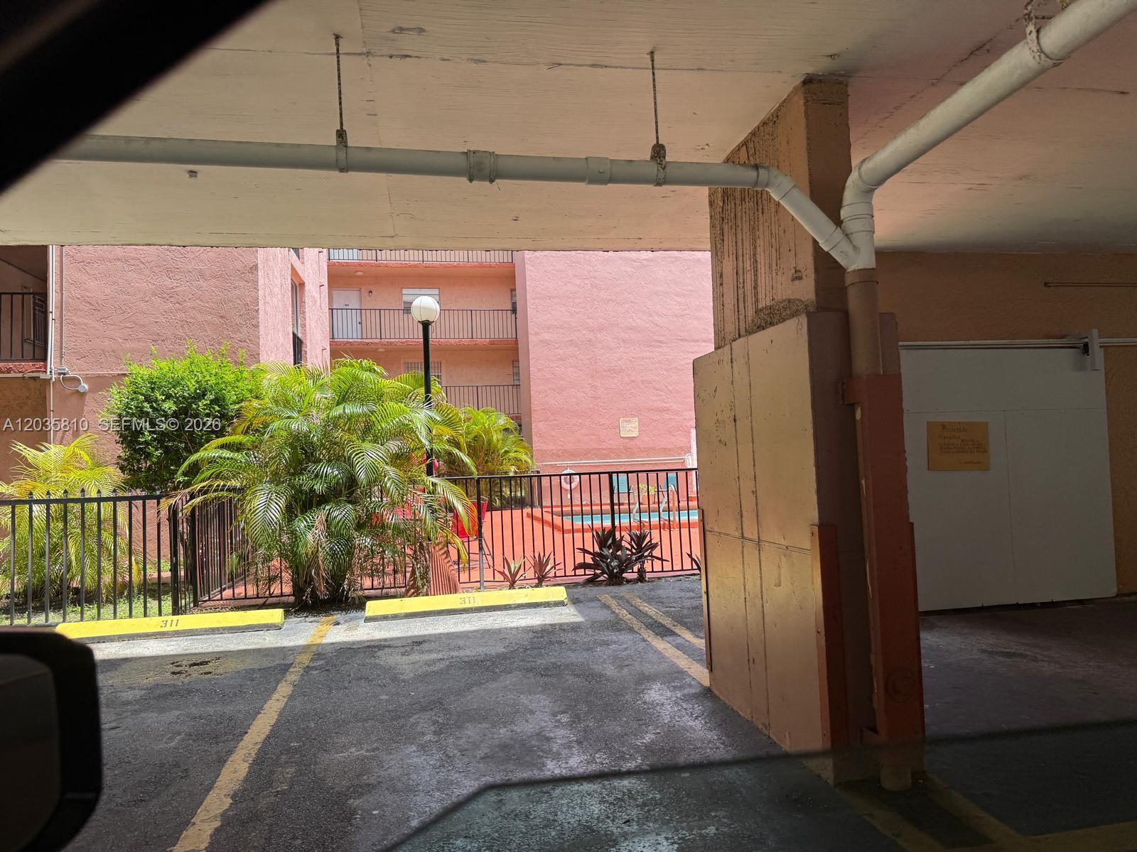 A covered parking space faces a fenced pool area with palm trees and a pink building — 6175 W 20th Ave # 210, Hialeah FL 33012, HIALEAH, FL
