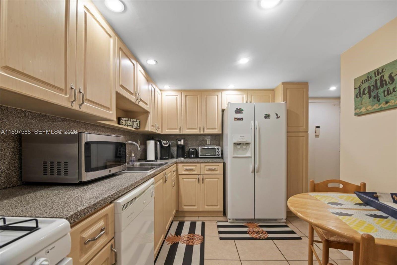 A kitchen features light wood cabinets, granite countertops, a white refrigerator, and a small dining table — 3140 S Ocean Dr # 207, Hallandale Beach FL 33009, HALLANDALE BEACH, FL