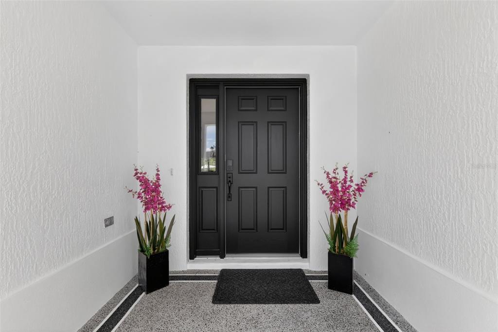 A black front door with a side window is flanked by two potted pink flowers — 6857 BAYSINGER PATH, THE VILLAGES, FL