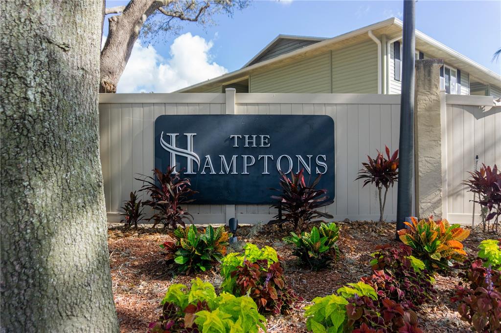 A sign for The Hamptons stands in front of a tan fence and landscaping — 502 GOLDEN RAINTREE PLACE, BRANDON, FL