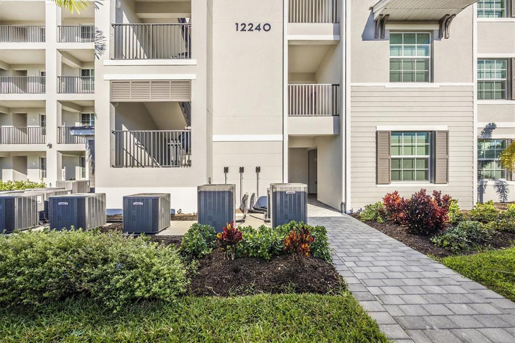 The exterior of a multi-story apartment building features a paved walkway and several air conditioning units — 12240 WELLEN GOLF STREET UNIT 207, VENICE, FL