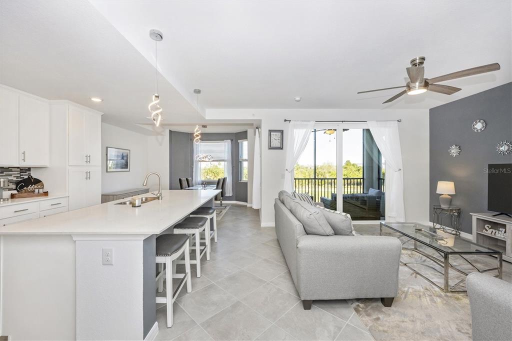 An open-concept kitchen and living area features a white island, gray sofa, and balcony access — 12240 WELLEN GOLF STREET UNIT 207, VENICE, FL