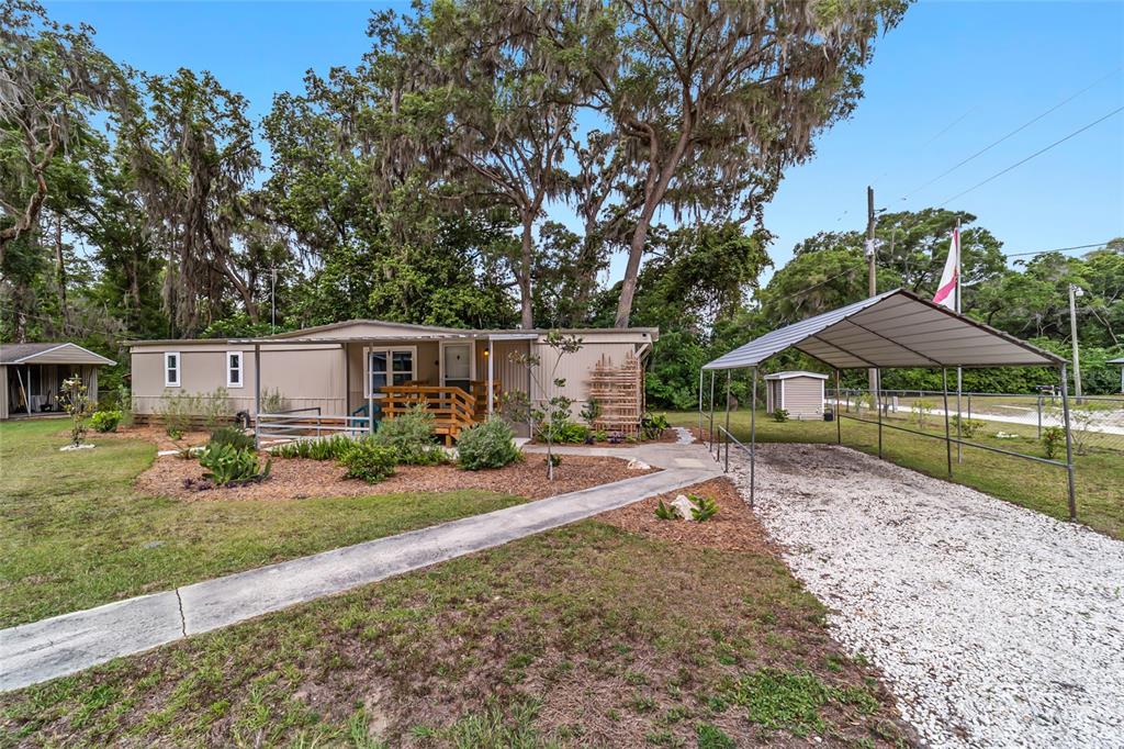 A single-story mobile home with a wooden porch, gravel carport, and mature trees in front — 3460 SE 131ST PLACE, BELLEVIEW, FL