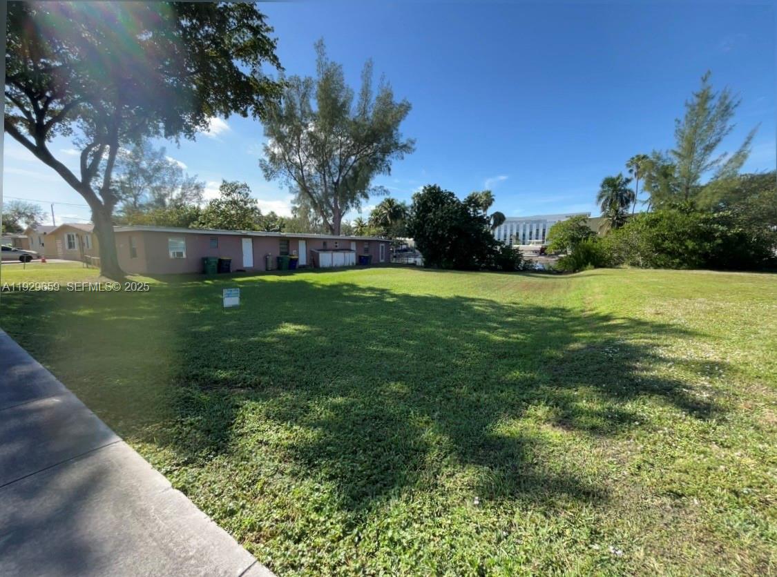 A grassy vacant lot sits adjacent to a single-story residential building under a clear sky — 157 NW 14th Way, Dania Beach FL 33004, DANIA BEACH, FL