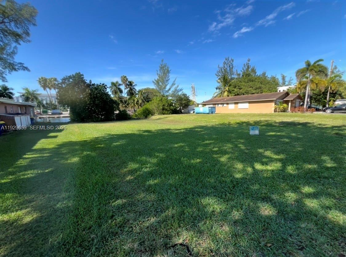 A grassy, vacant residential lot features a small sign and mature trees under a blue sky — 157 NW 14th Way, Dania Beach FL 33004, DANIA BEACH, FL