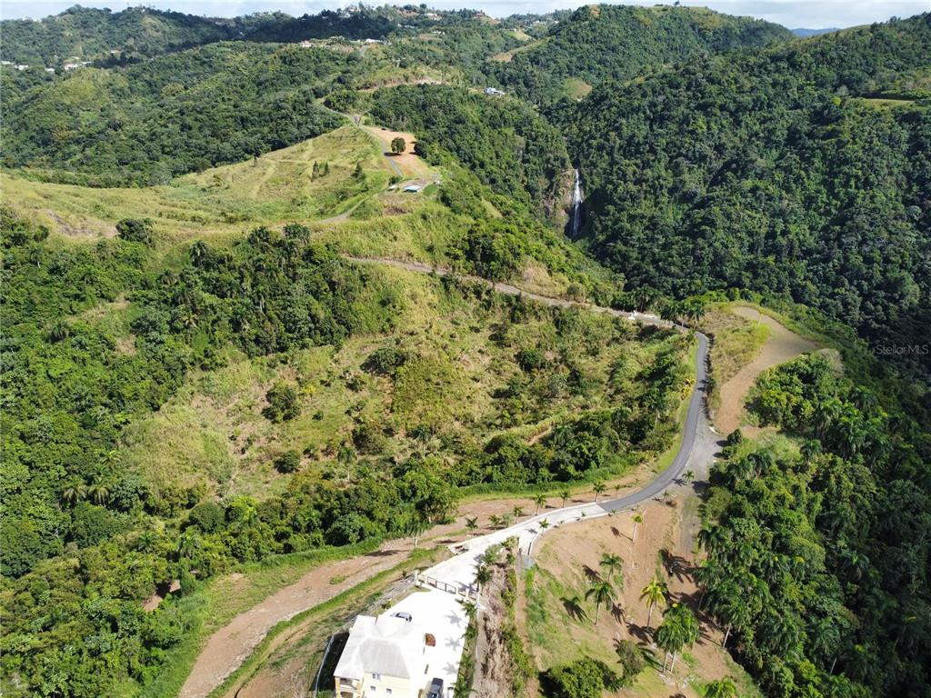 An aerial view shows a house with a concrete driveway nestled in a mountainous landscape — PR-155 KM 34.0 BARRIO PERCHAS, MOROVIS, PR