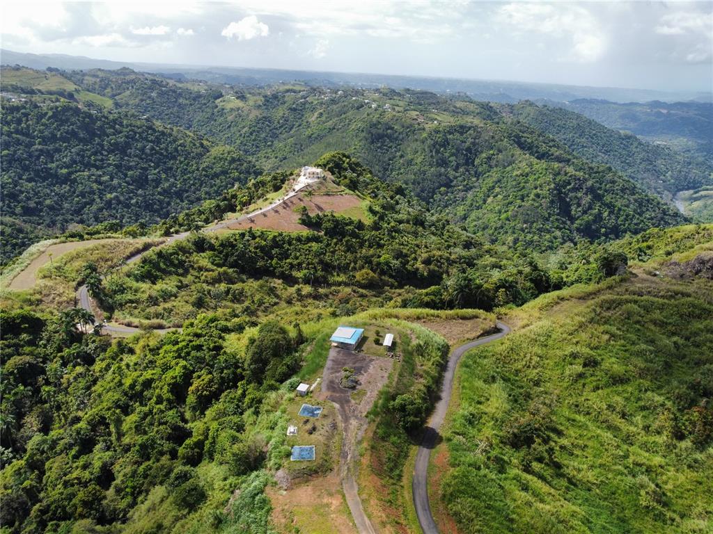 An aerial view shows a winding road leading to a hilltop house amidst mountainous terrain — PR-155 KM 34.0 BARRIO PERCHAS, MOROVIS, PR
