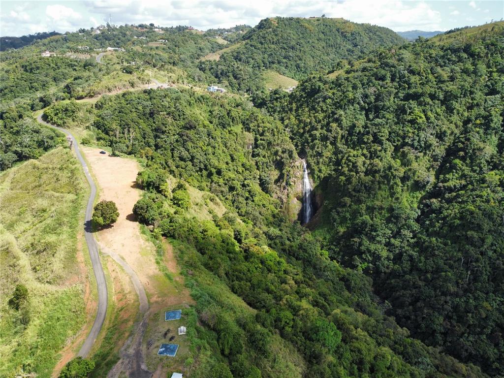 An aerial view shows a waterfall nestled in a lush, green, mountainous landscape with a road — PR-155 KM 34.0 BARRIO PERCHAS, MOROVIS, PR