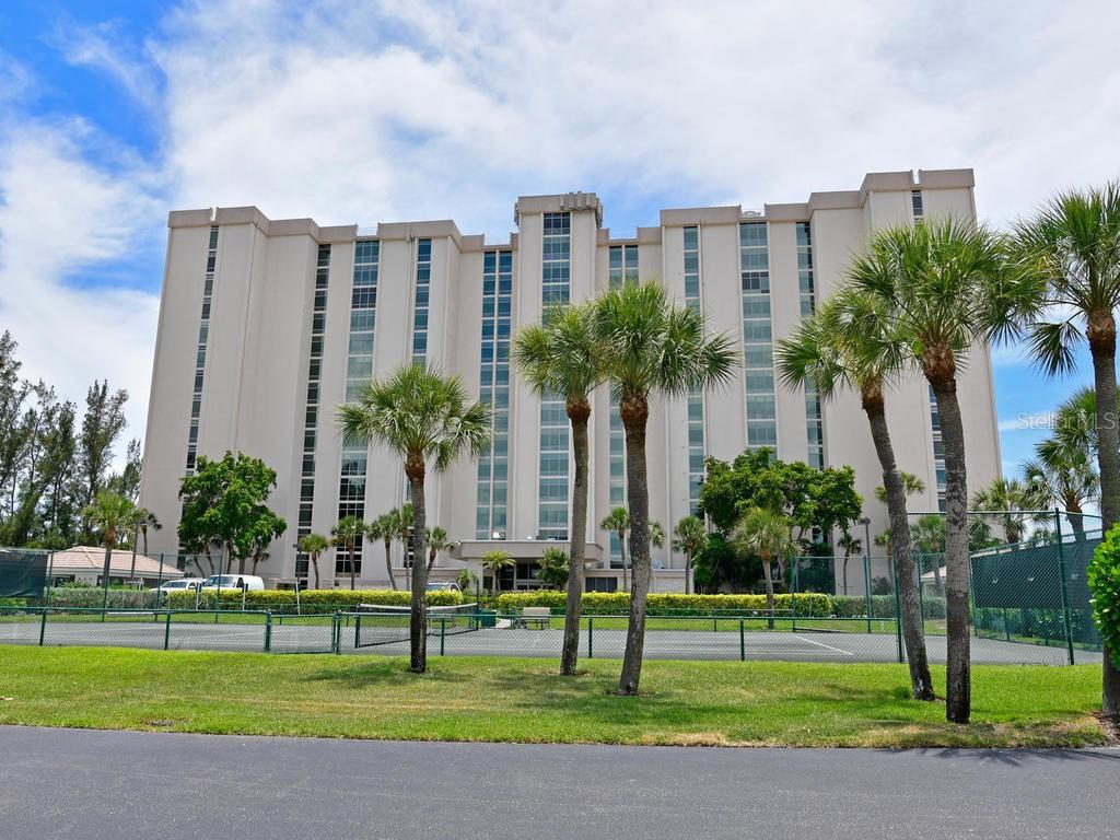 A multi-story beige apartment building stands behind a fenced tennis court with palm trees — 2425 GULF OF MEXICO DRIVE UNIT 7F, LONGBOAT KEY, FL