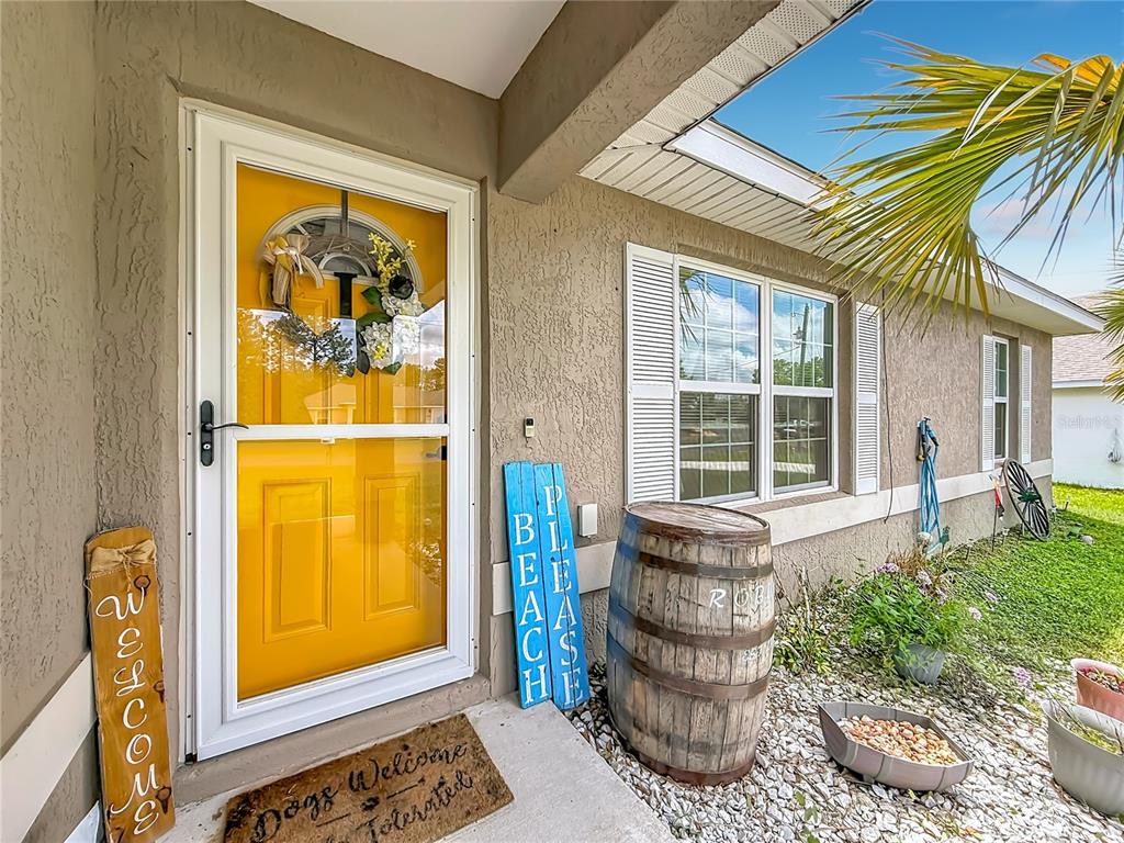 A bright yellow front door with a wreath sits beside a wooden barrel and signs — 6050 SW 155TH STREET ROAD, OCALA, FL