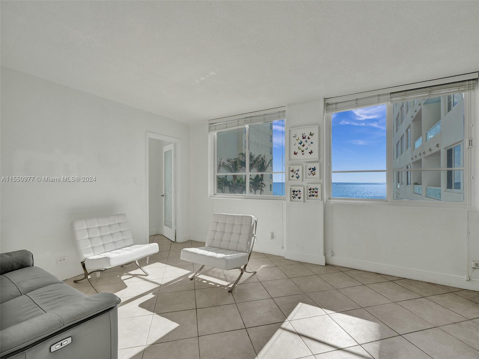 A bright living room features white leather chairs, tile floors, and ocean views through windows — 5005 Collins Ave # 725, Miami Beach FL 33140, MIAMI BEACH, FL