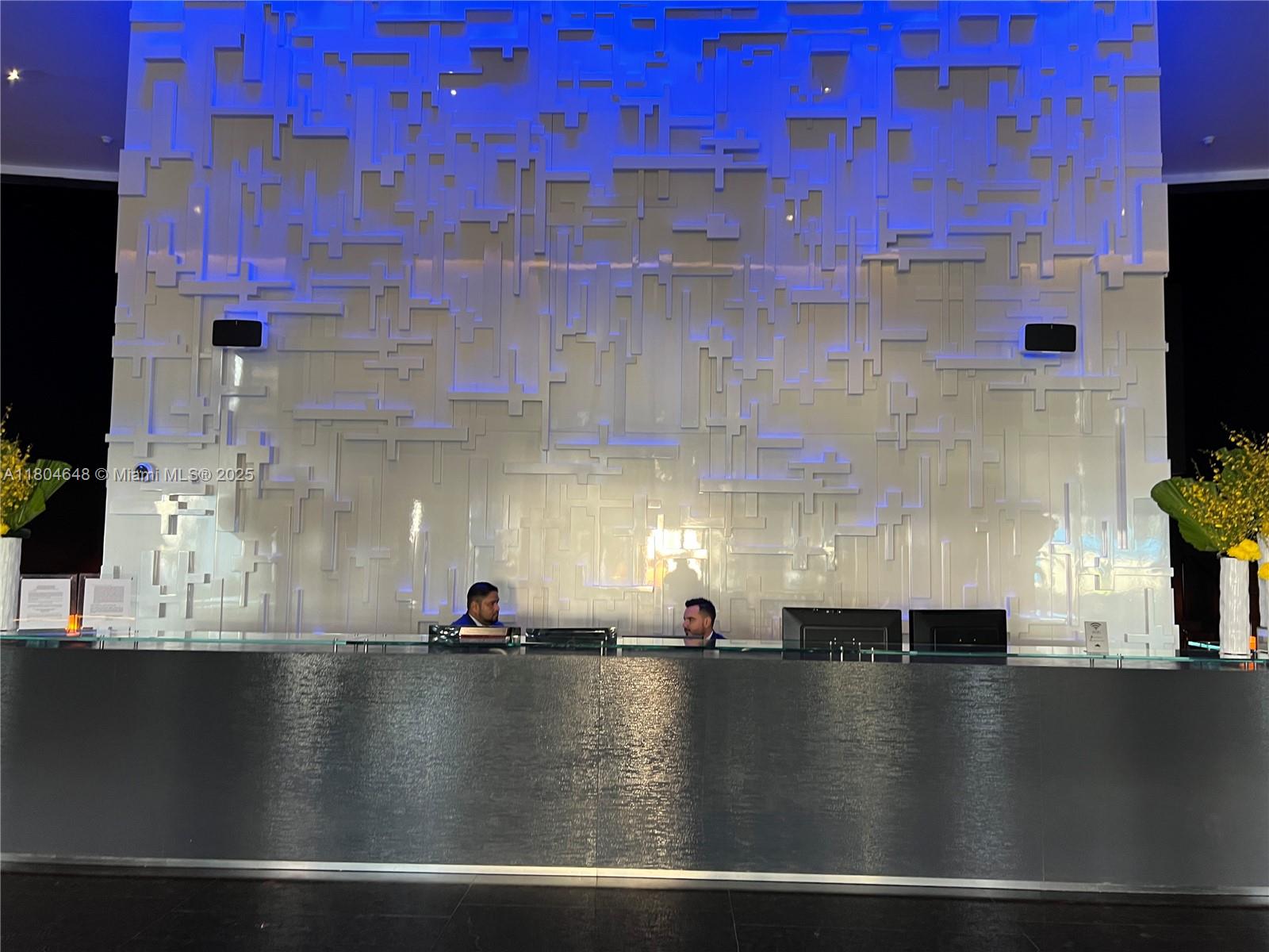 A modern reception desk features a textured gray front and a large, illuminated geometric wall — 1800 S Ocean Dr # 2006, Hallandale Beach FL 33009, HALLANDALE BEACH, FL