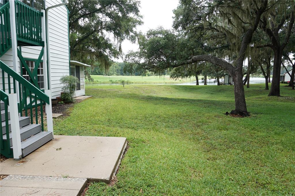 A view of a grassy backyard with a pond, large trees, and a building exterior — 7945 MIDWAY DRIVE TERRACE UNIT H201, OCALA, FL