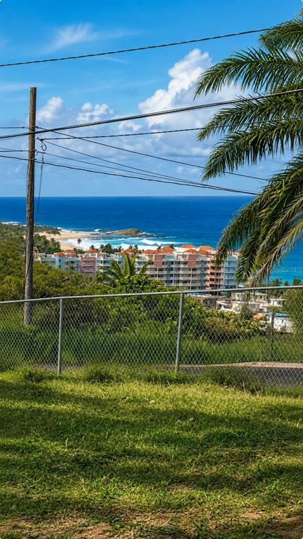 A chain-link fence overlooks a grassy hill, a coastal resort, and the blue ocean — CALLE PASCUA URB. MANUEL CORCHADO, ISABELA, PR