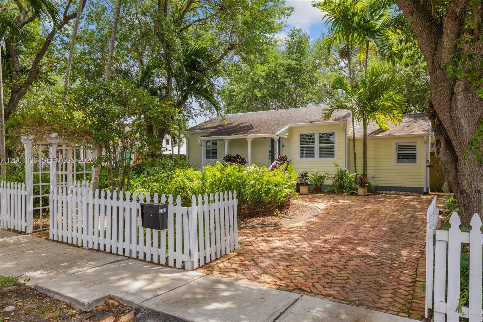 A yellow house with a brick driveway and white picket fence sits behind lush landscaping — 6382 SW 38th St, Miami FL 33155, MIAMI, FL