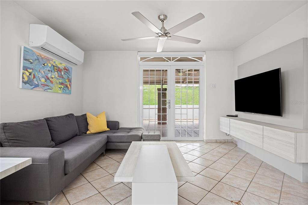 A living room features a grey sectional sofa, a wall-mounted television, and tiled flooring — Calle 6 H17 TINTILLO GARDENS, GUAYNABO, PR