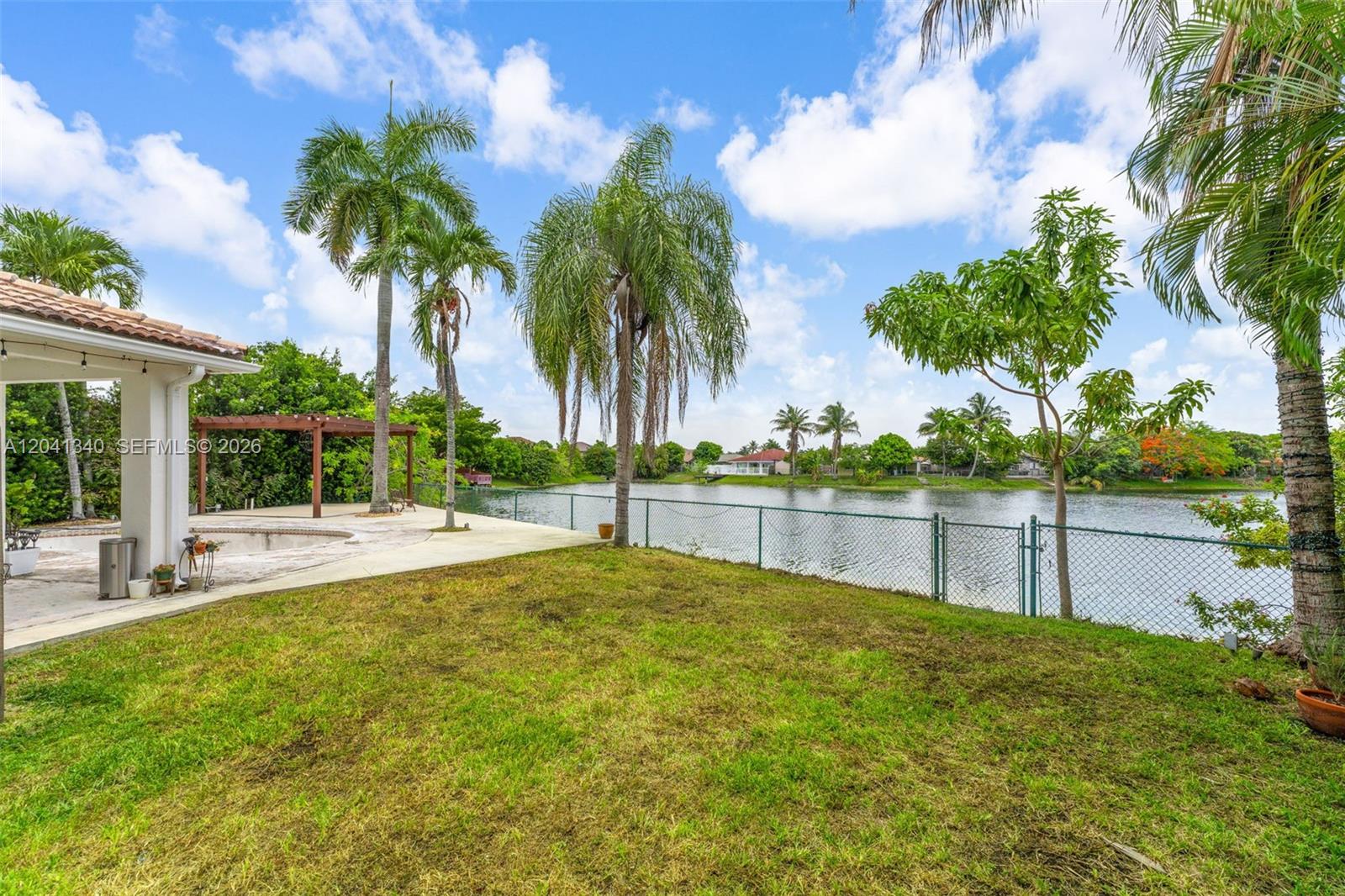 A grassy backyard features a patio, palm trees, and a chain-link fence overlooking a lake — 16335 SW 81 St, Miami FL 33193, MIAMI, FL