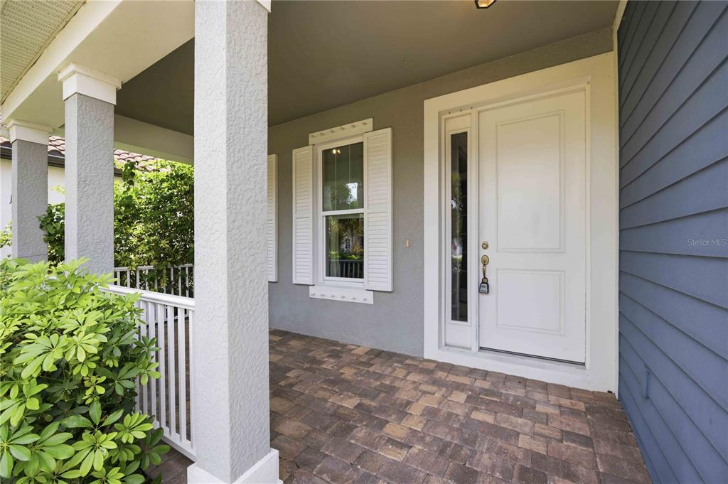 A covered front porch features a white door, a window with shutters, and paver flooring — 11516 RENAISSANCE BOULEVARD, VENICE, FL